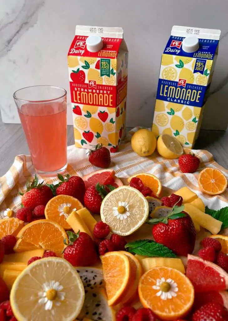 A fruit board with 2 cartons & a glass of AE lemonade in the background. 